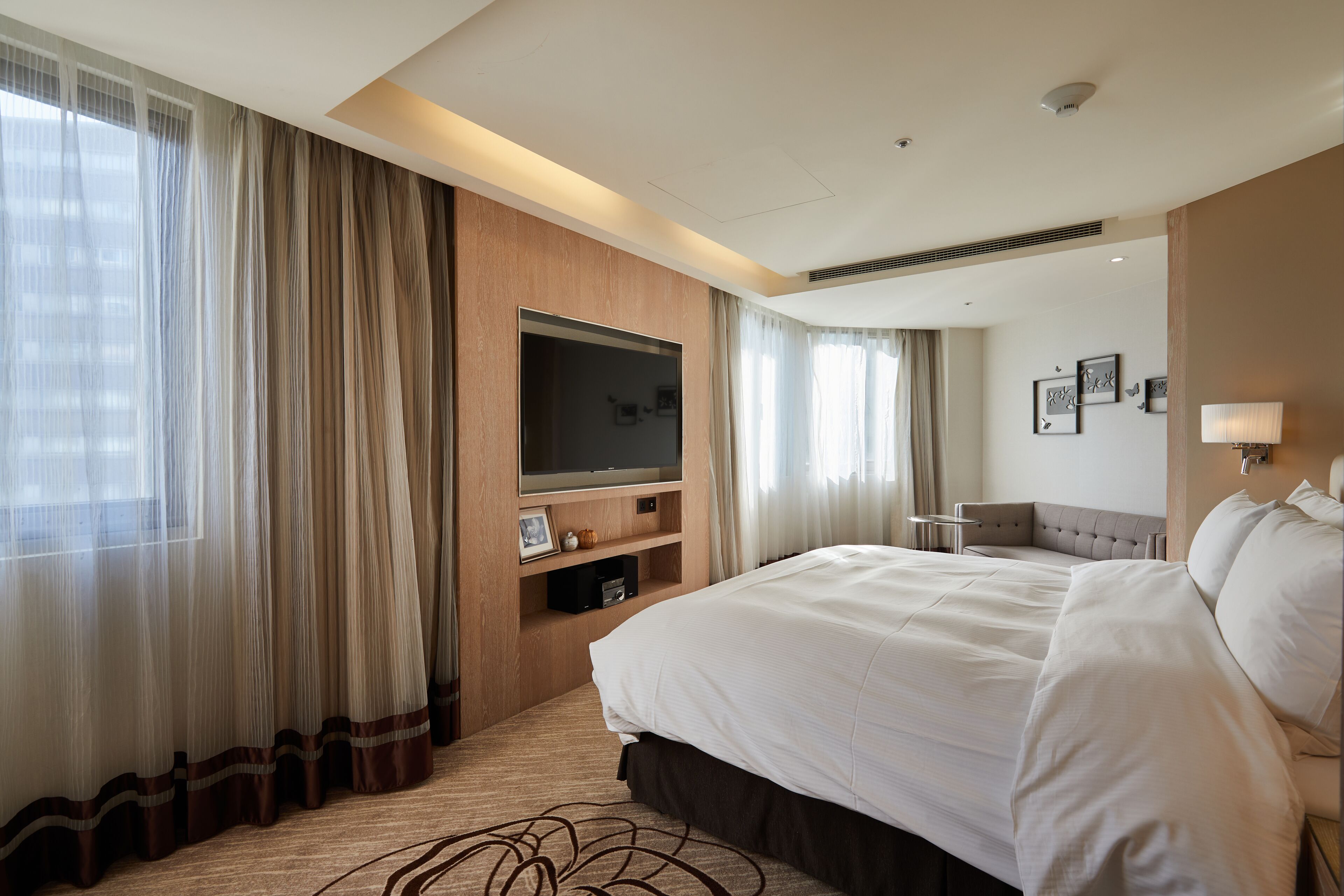 A hotel room with a large bed, a flat-screen TV mounted on the wall, a seating area with a chair, and a large window with sheer curtains.