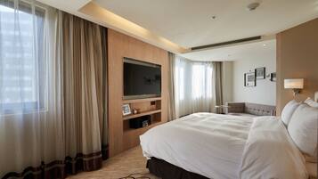 A hotel room with a large bed, a flat-screen TV mounted on the wall, a seating area with a chair, and a large window with sheer curtains.