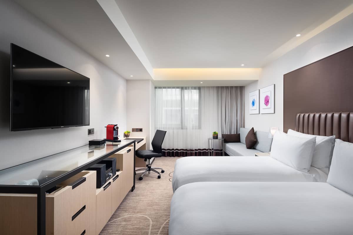 A hotel room with two beds, a desk with a TV, a chair, and a small table with a plant.
