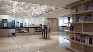 Eine moderne Hotellobby mit einer zentralen Gastronomie, einer Rezeption und einer Bücherwand mit Büchern und Zeitschriften.