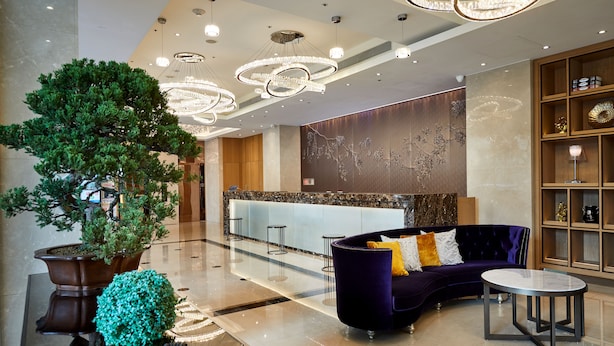 Eine moderne Lobby mit einer Rezeption, einem großen Kübelbaum, einem violetten Sofa mit rundem Tisch und einer Kristallleuchter.