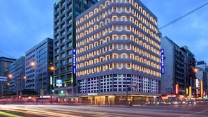 Ein mehrstöckiges Hotel mit modernem Design, das durch seine exponierte Ecklage, beleuchtete Fenster und Beschilderung auffällt.