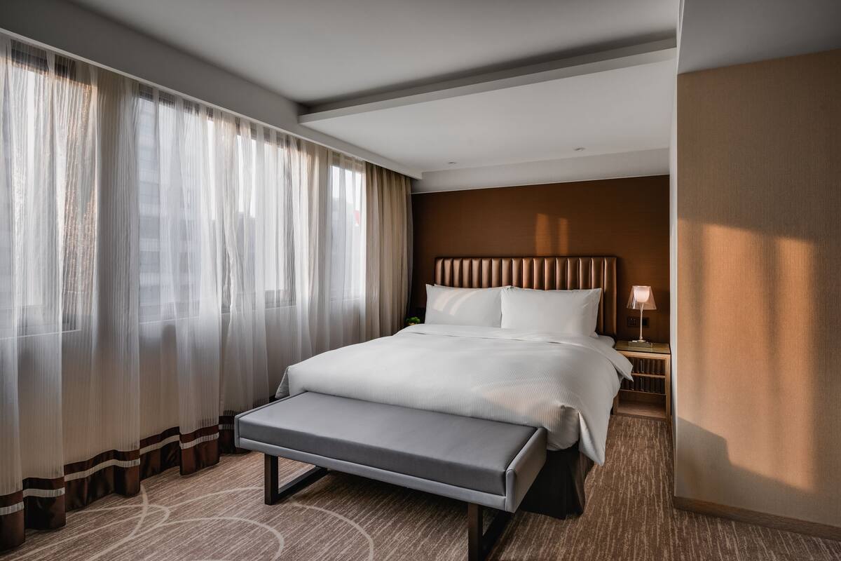 A hotel room with a large bed, a bench, a nightstand, and a window with sheer curtains.