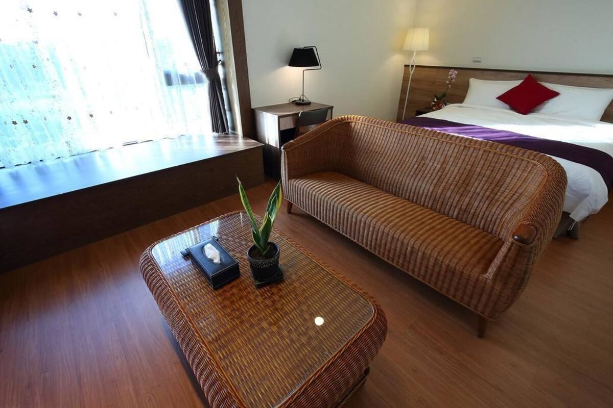 A hotel room with a wicker sofa, a small table with a plant, a bedside lamp, and a bed with white and red bedding.