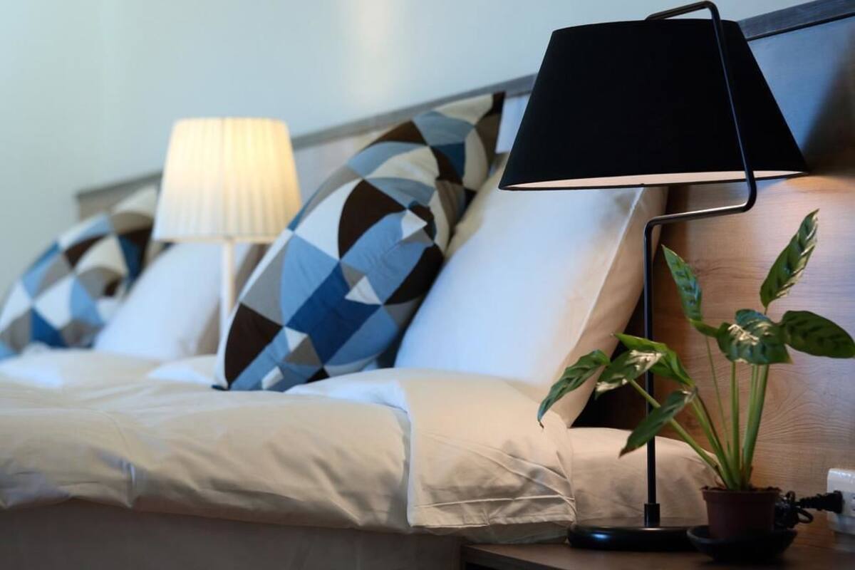 A neatly made bed with a white comforter, a black lamp on a bedside table, and a potted plant.