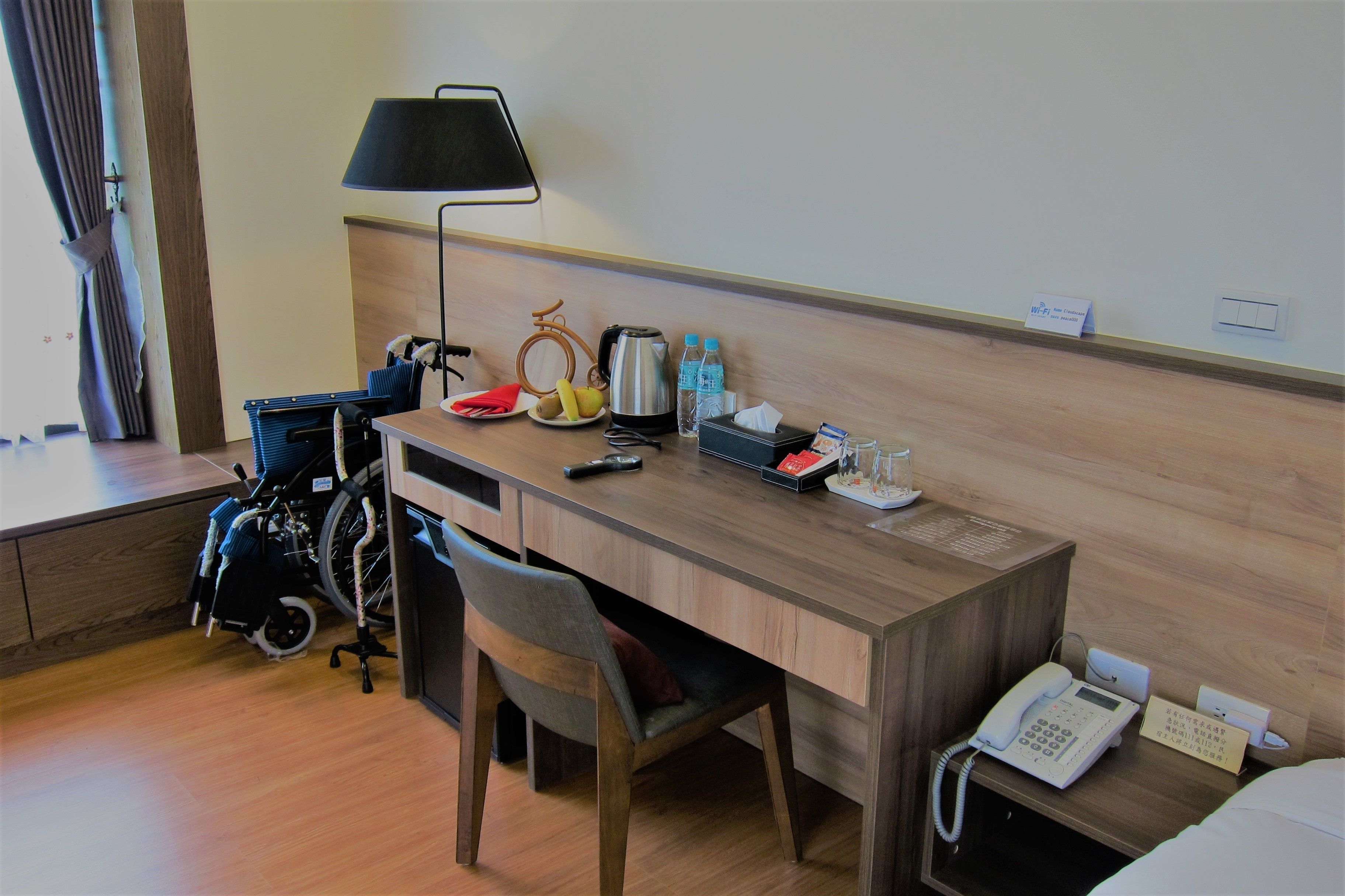 A hotel room with a wooden desk, a chair, a bed, a bedside phone, a lamp, a coffee maker, a plate with fruit, a water bottle, a remote control, and a wheelchair.
