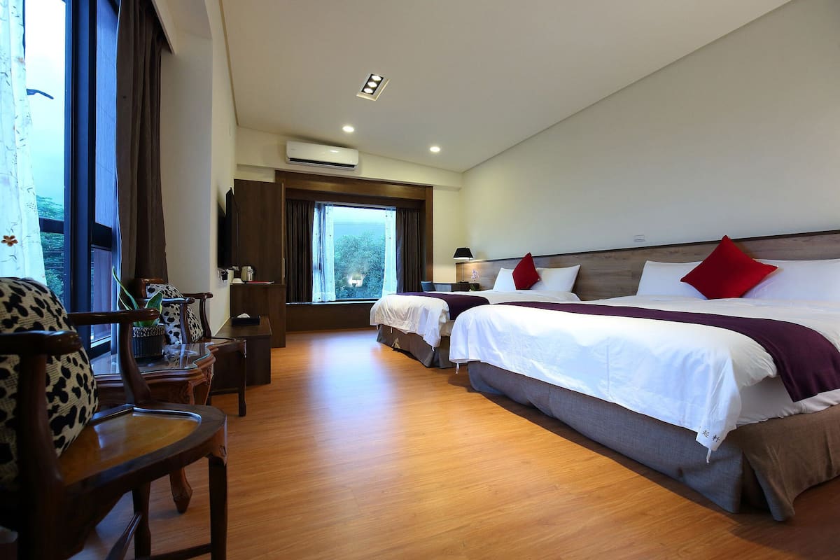 A hotel room with two beds, wooden flooring, a desk with a chair, a window with curtains, and an air conditioning unit.
