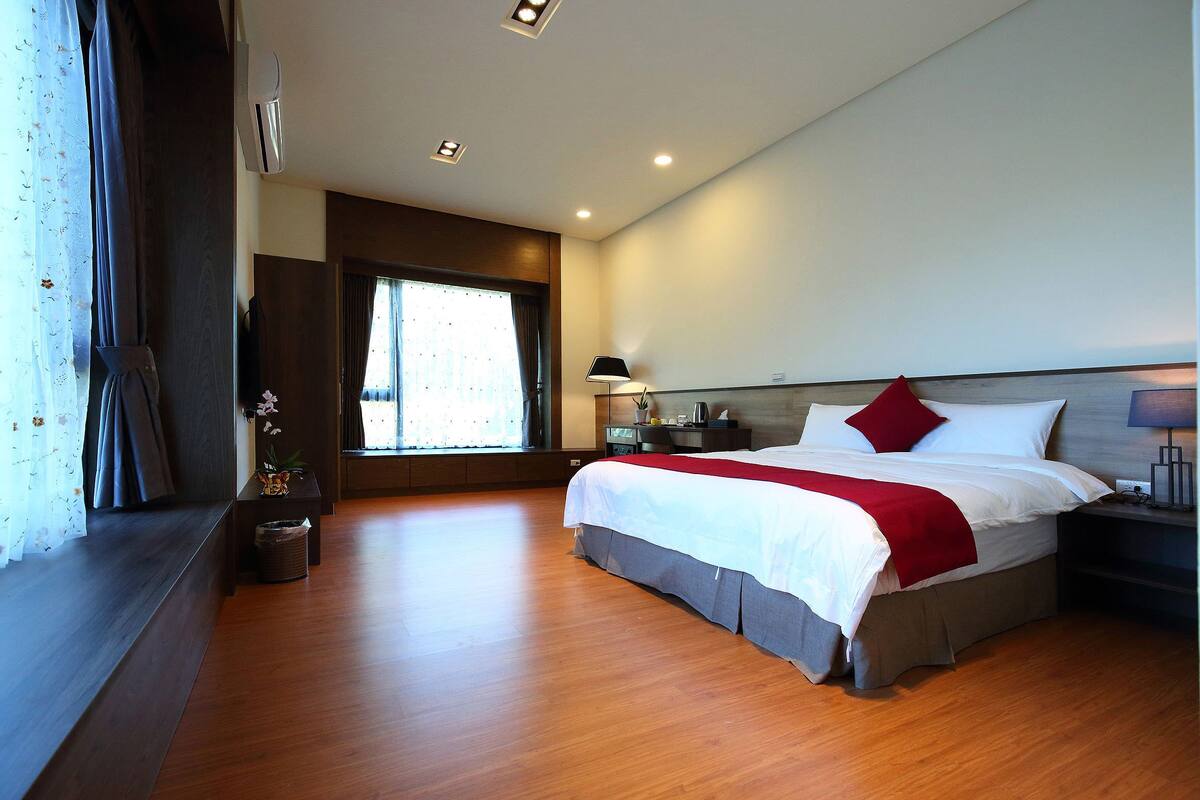 A modern hotel room with a large bed, a bedside table, a nightstand, a window with curtains, and a ceiling light.