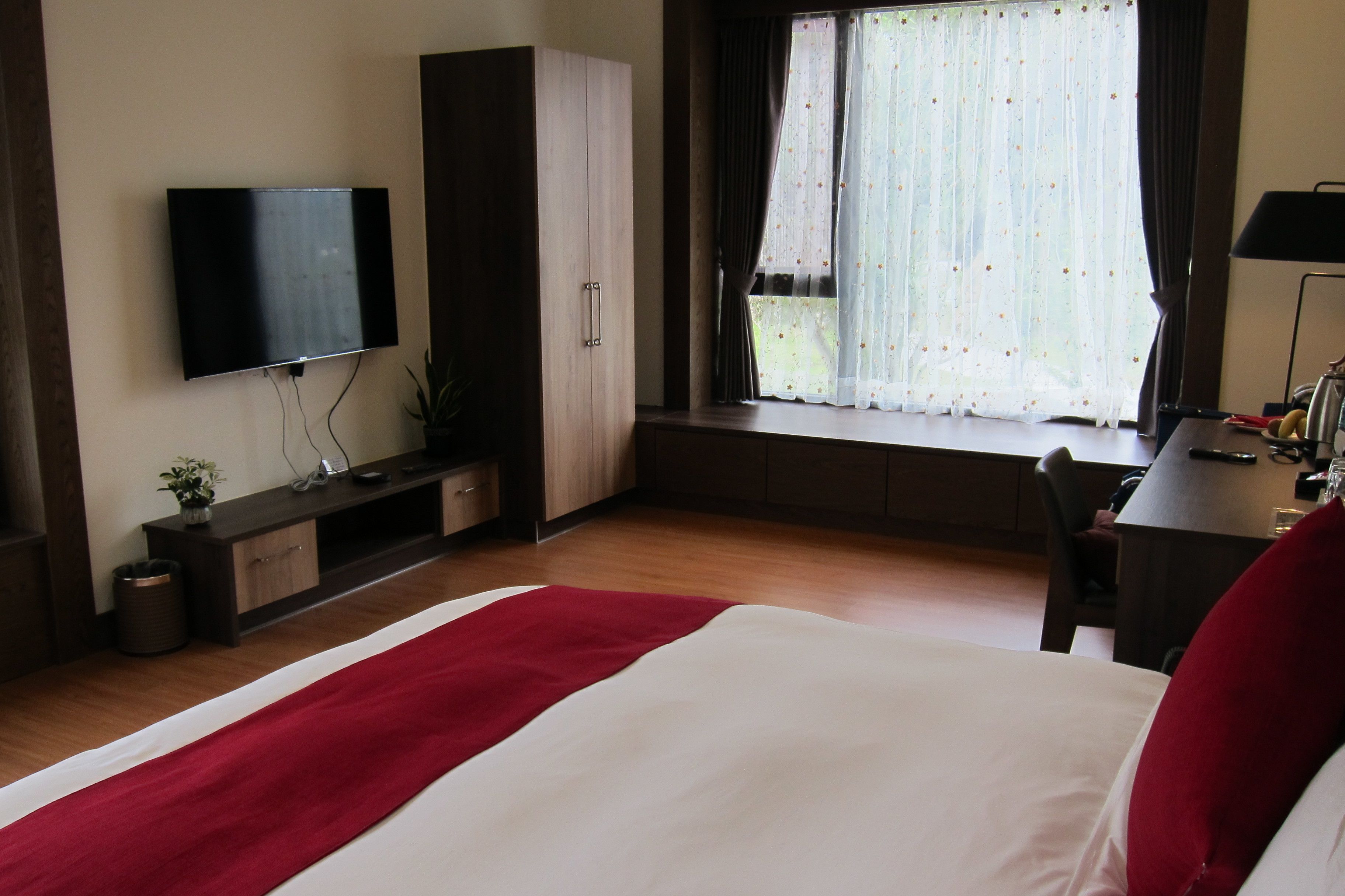 A hotel room with a bed, a TV mounted on the wall, a desk with a lamp, and a window with curtains.