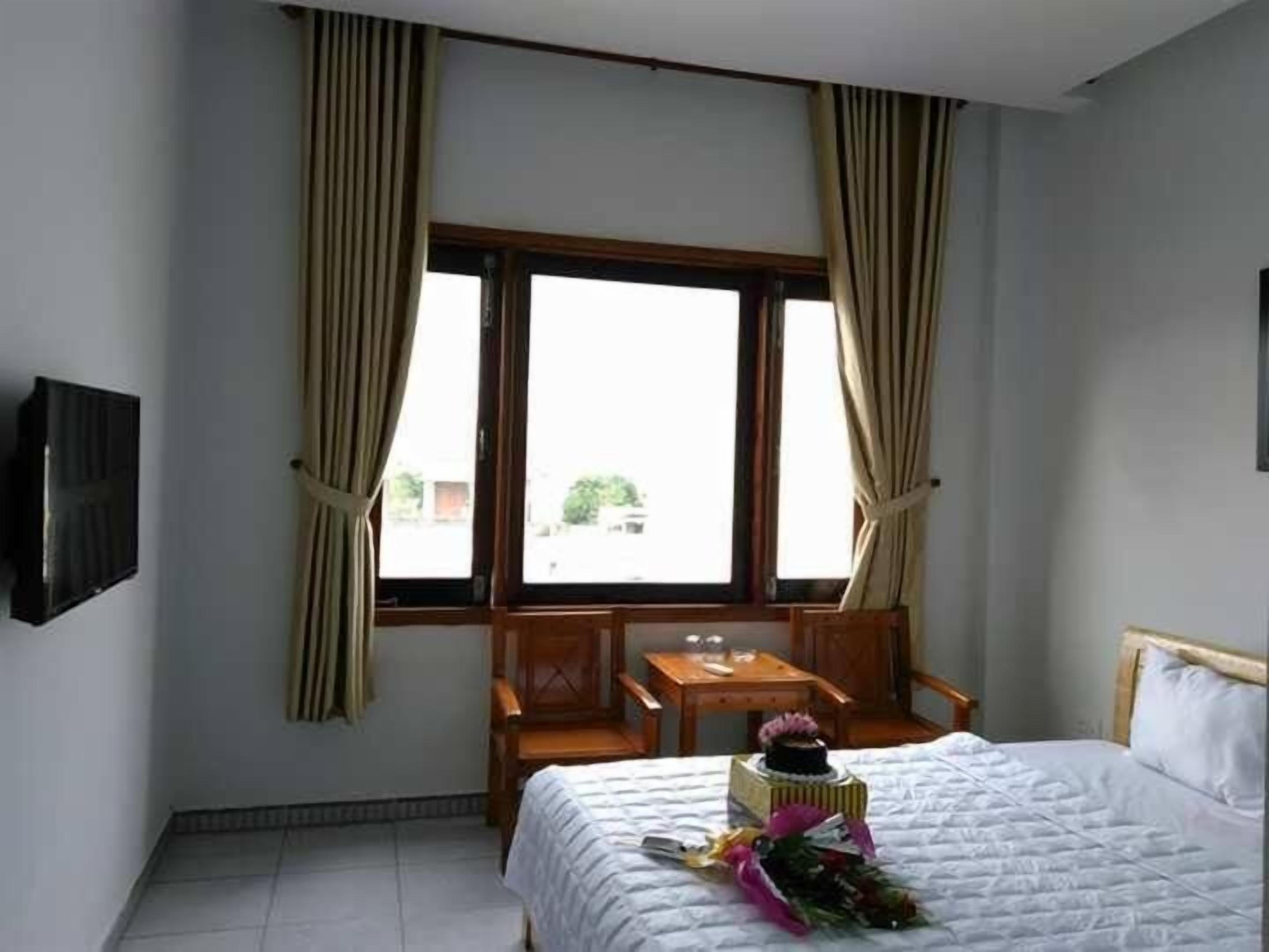 A hotel room with a bed, a small table with a vase of flowers, a chair, and a window with curtains.
