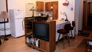 A compact kitchen with wooden cabinets, a refrigerator, a stove, and a dining area with two chairs. - Small cabin in the woods Peaceful retreat (East Bernstadt)