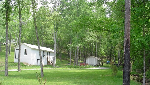 A white cabin with a small porch, surrounded by a wooded area with tall trees and a grassy field. - Small cabin in the woods Peaceful retreat (East Bernstadt)