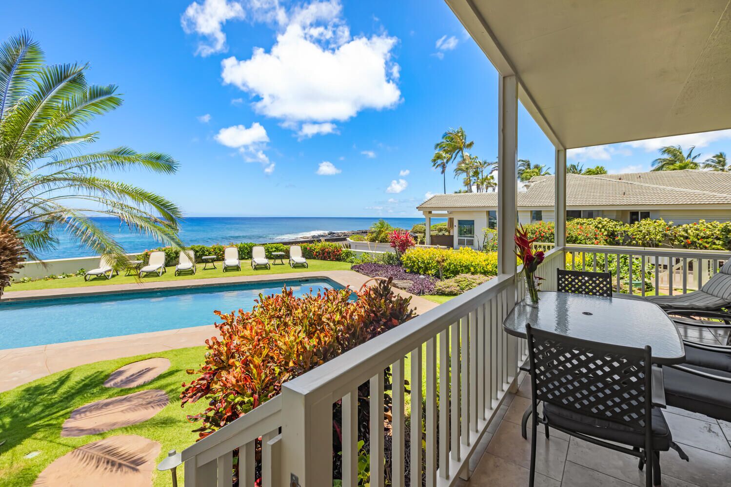 A poolside area with a view of the ocean, lounge chairs, and a covered patio with a table and chairs.