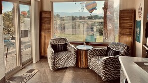 A room with two patterned armchairs, a small round table, and a view of a hot air balloon and landscape through large windows. - Golf Course, Mnt Views, Balloons, King Bed, Veranda, Yard, 3 BD, WIFI, BBQ, WD (Albuquerque)