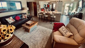 A living room with a sofa, coffee table, and a chair with a 'HOME' pillow. There is a kitchen area with bar stools and a refrigerator. - Golf Course, Mnt Views, Balloons, King Bed, Veranda, Yard, 3 BD, WIFI, BBQ, WD (Albuquerque)