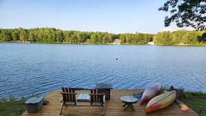 A wooden deck with two chairs and a table, a kayak, and a paddle on the shore of a lake.