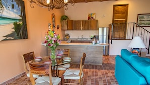 Comedor con mesa de vidrio, sillas de madera y un florero. Se observa una cocina con gabinetes de madera y un lavamanos. Un sofá azul es visible al fondo.