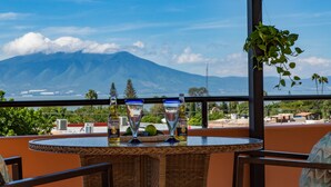 Una terraza con vista a la montaña, una mesa con dos vasos y una botella, y mobiliario de mimbre.