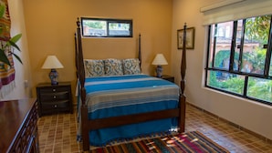 Un dormitorio con cama con dosel, dos mesitas de noche con lámparas, una ventana con vista a follaje y una alfombra estampada.