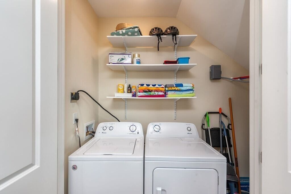 A laundry room with two front-loading washers, shelves above holding various items, and a mop on the right.