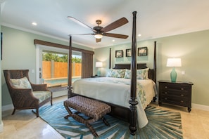 A bedroom with a four-poster bed, a ceiling fan, a nightstand with a lamp, and a view of the outdoors.