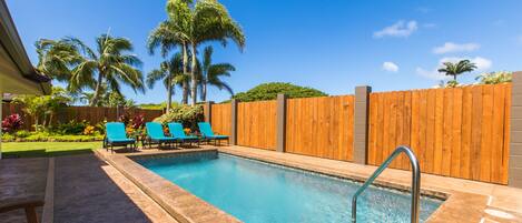 A swimming pool with a wooden fence, blue lounge chairs, and palm trees in the background.