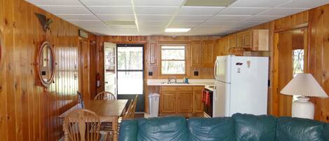 Interior de una cabaña con revestimiento de madera, un comedor con sillas, un sofá, una cocina con refrigerador y una ventana.