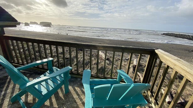 Una terraza de madera con sillas turquesas que da a una playa con olas y rocas.