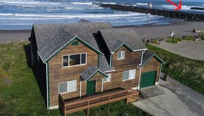 A wooden house with a green roof and a deck, located near a rocky shoreline with the Coquille River visible in the background. - Walk out onto the beach! (Bandon)