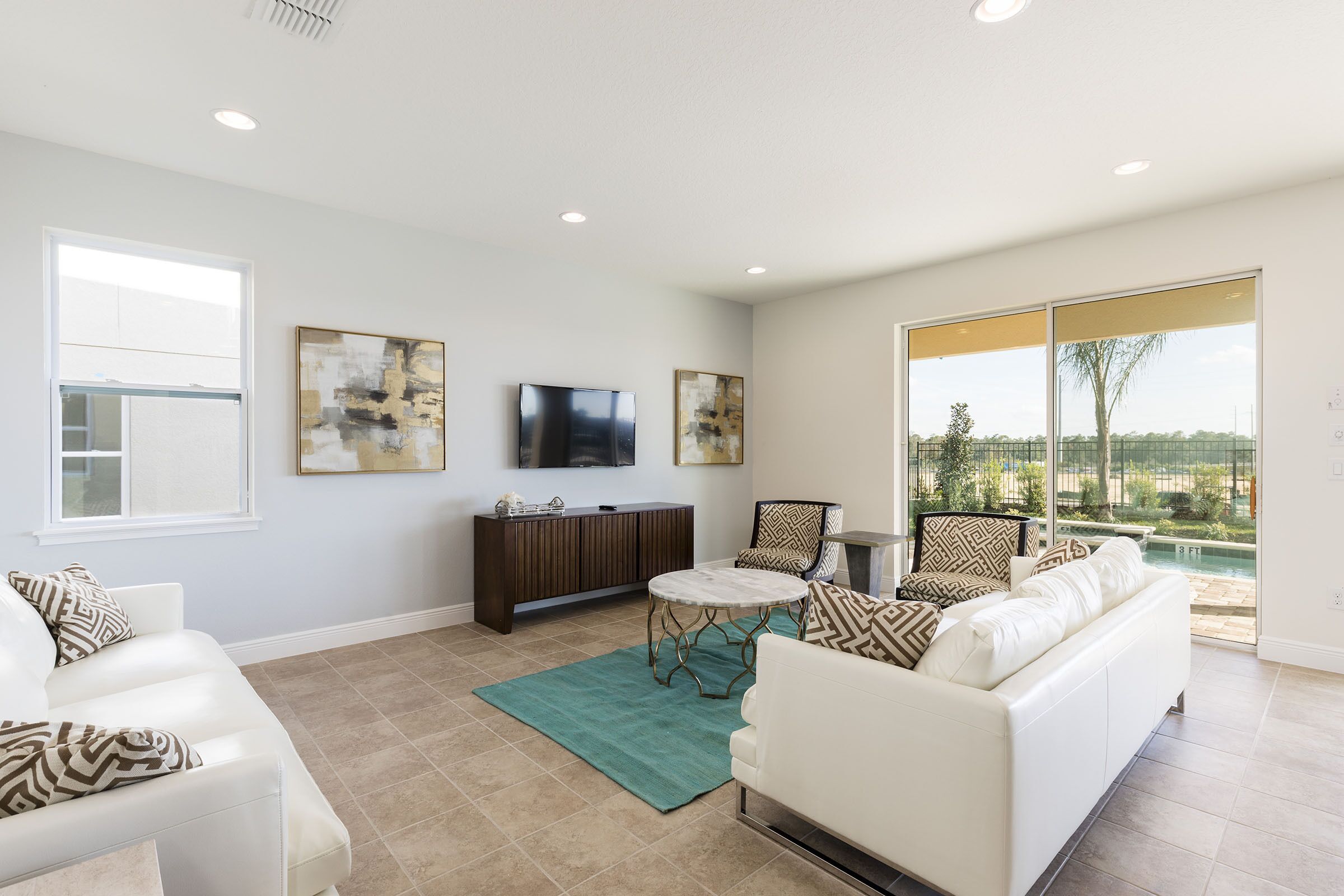A modern living room with a white sofa, a coffee table, and a TV on a wall. There is a large window with a view of a pool and greenery.