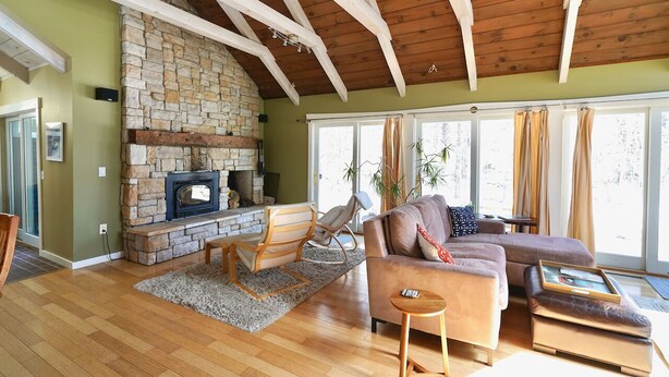 A living room with a stone fireplace, wooden beams, a sofa, and a large window with curtains. - Perfectly located on the Mountain rd between Stowe village and Stowe Mountain (Stowe)