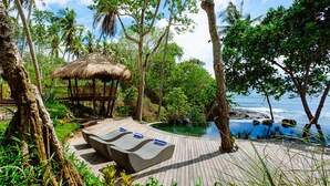 Une terrasse en bois avec des chaises longues et une structure à toit de chaume, offrant une vue sur une piscine et l’océan.