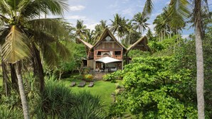 Une maison en bois à deux étages, dotée d’un toit de chaume, entourée d’une végétation luxuriante et de palmiers.