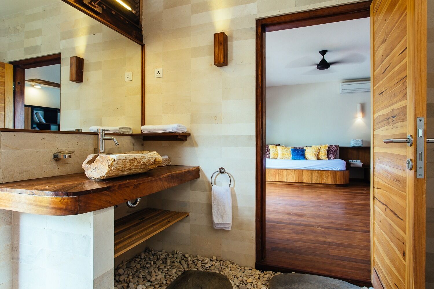 Un baño con tocador de madera, un espejo grande y una cama visible a través de una puerta abierta.