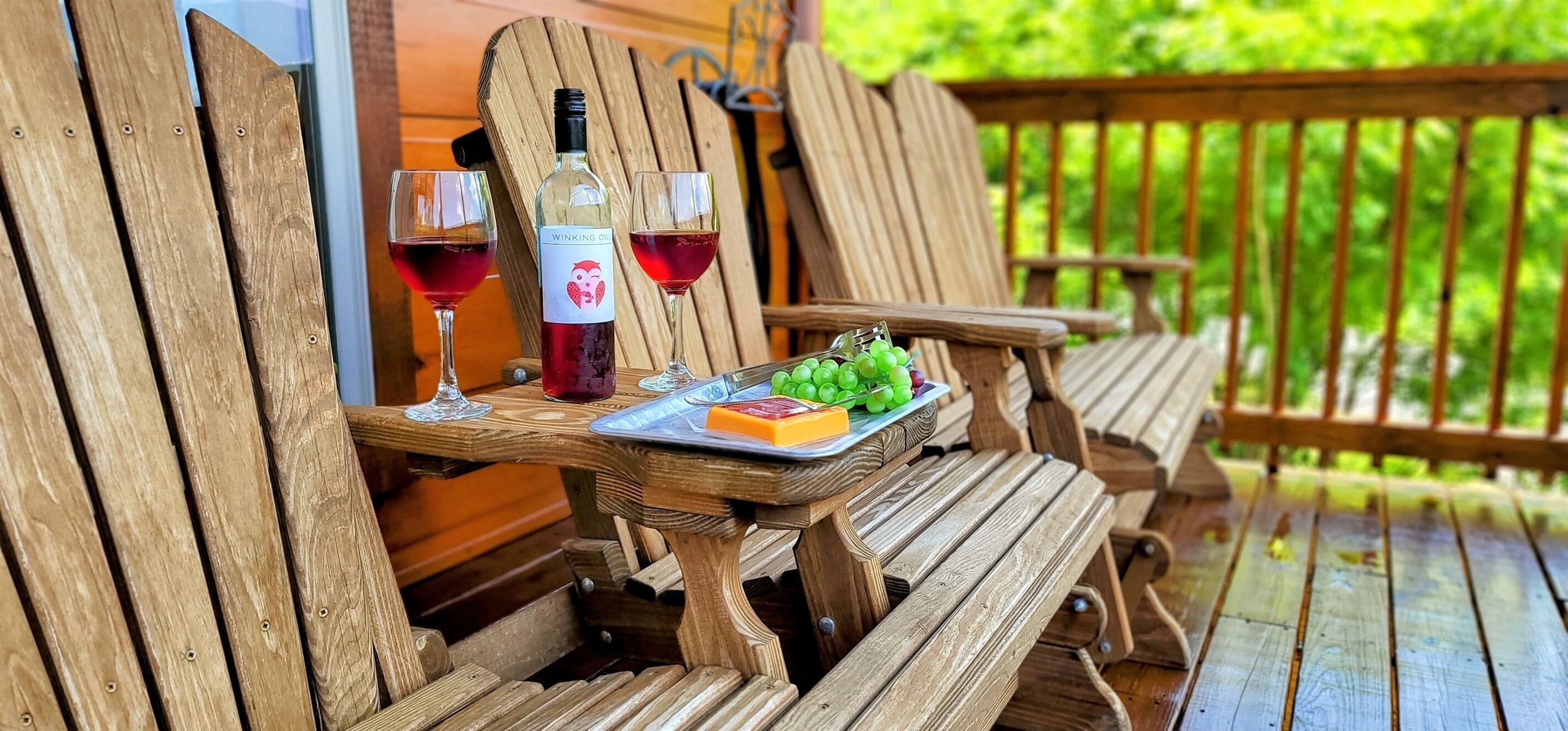 Une terrasse en bois avec une table dressée pour deux personnes, agrémentée d’une bouteille de vin, de deux verres, d’un plateau de fromages et d’une assiette de raisins.
