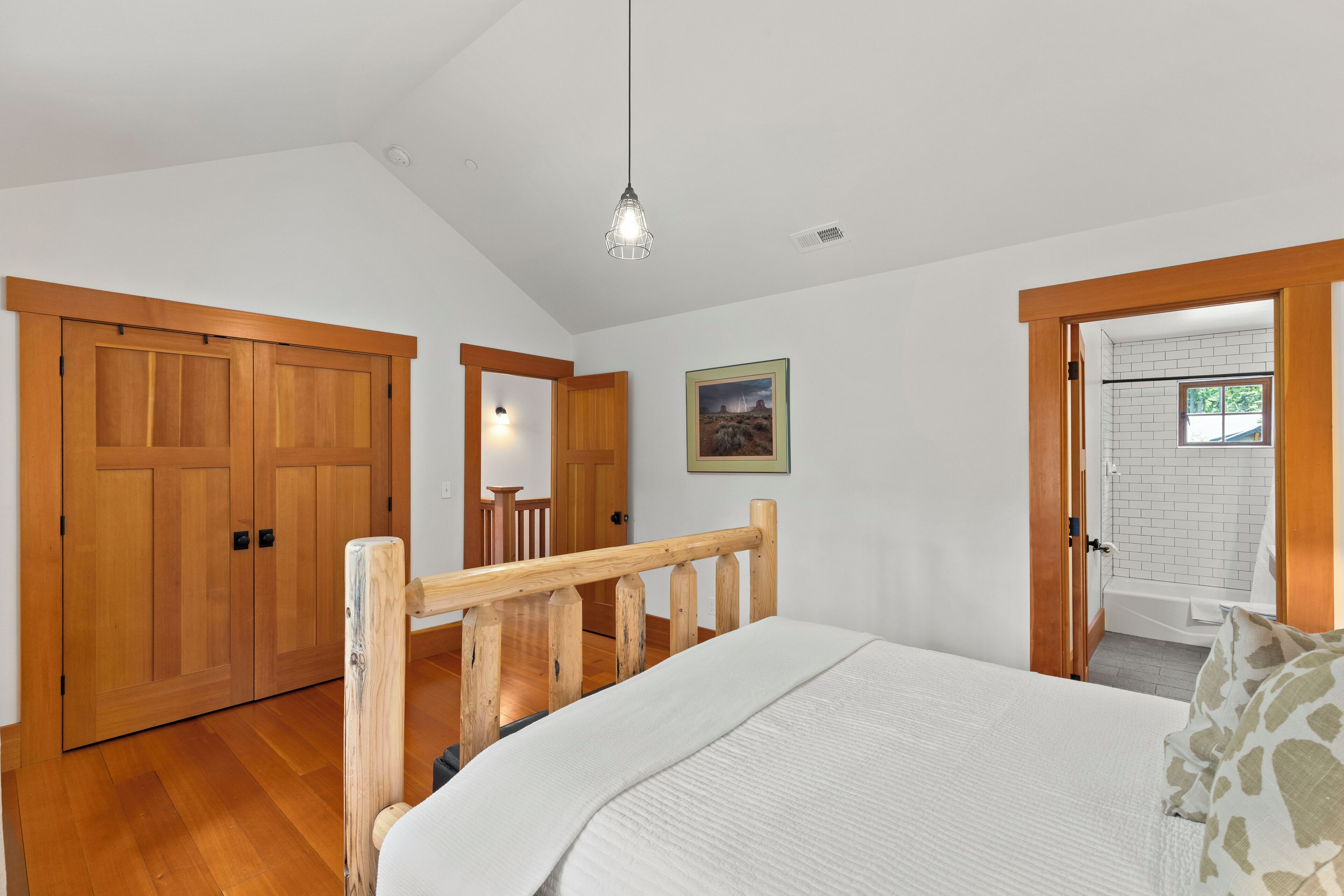 Ein Schlafzimmer mit Holzbettgestell, einer weißen Matratze, einem Holz-Kopfende, einer Hängeleuchte und Blick auf ein Badezimmer mit Badewanne und Fenster.