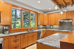 A kitchen with wooden cabinets, granite countertops, a stainless steel dishwasher, and a large window with a view of trees. - Suncadia Retreat • half acre • 3 porches • Quiet Cul-de-Sac • Fire pit • Bikes (Cle Elum)