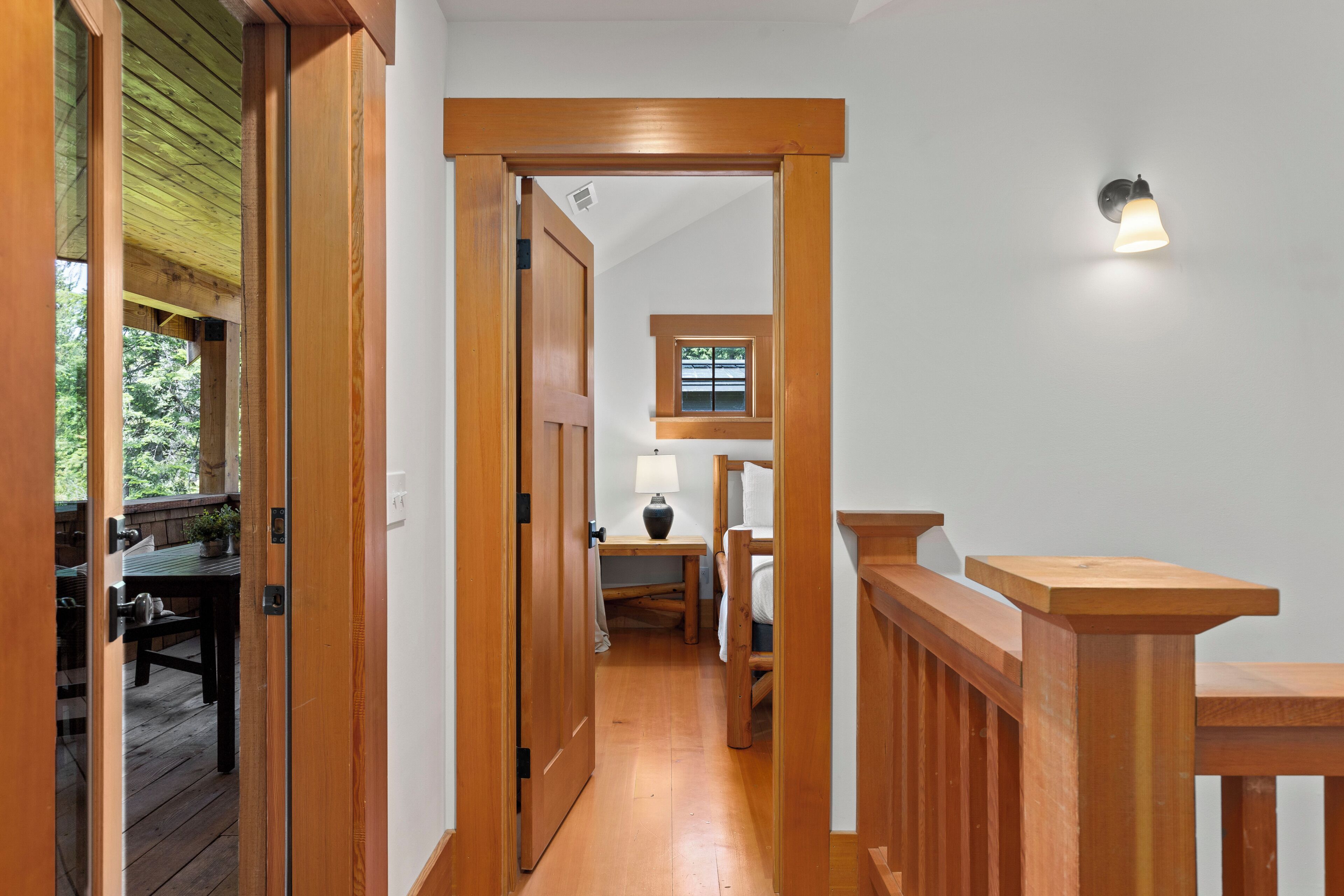A wooden hallway with a door leading to a room, a small table with a lamp, and a window with a view of greenery.