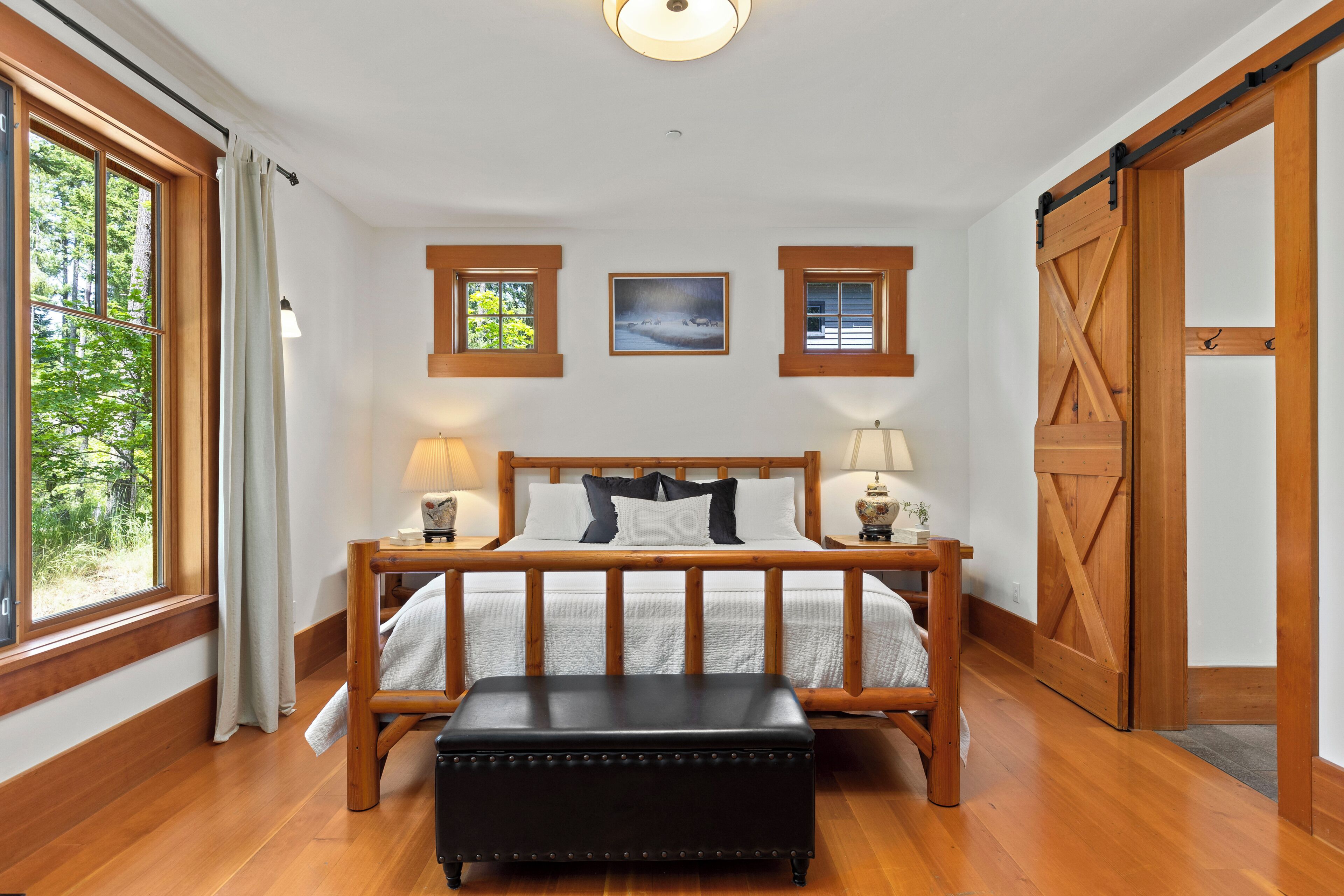 A bedroom with a wooden bed, two bedside lamps, a bench, a large window, and a sliding barn door.