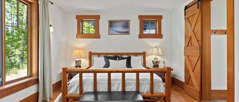 A bedroom with a wooden bed, two bedside lamps, a bench, a large window, and a sliding barn door.