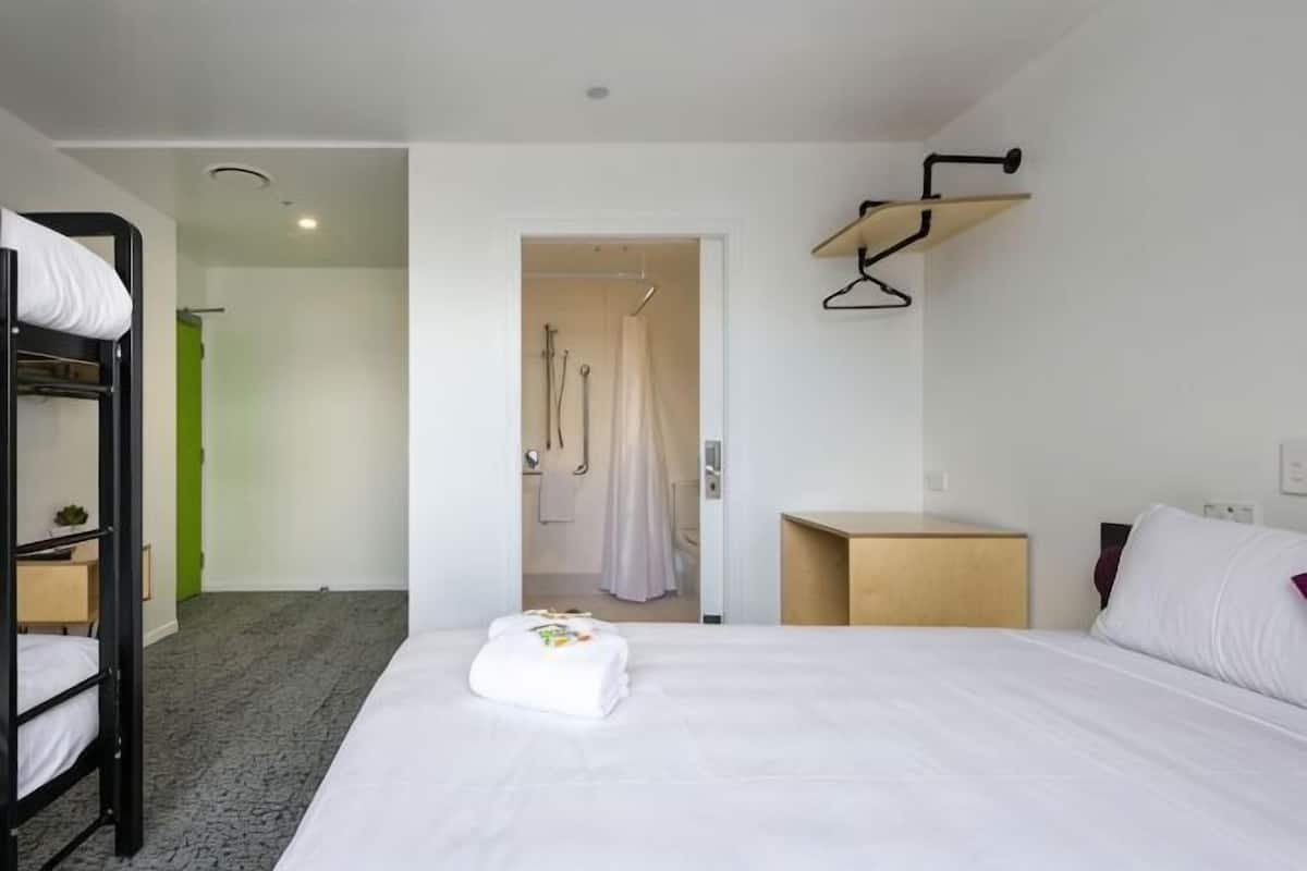 Ein Hotelzimmer mit Etagenbett, einem Einzelbett, einem Nachttisch aus Holz, einer Hängeleuchte und einem sichtbaren Duschbereich durch eine geöffnete Tür.