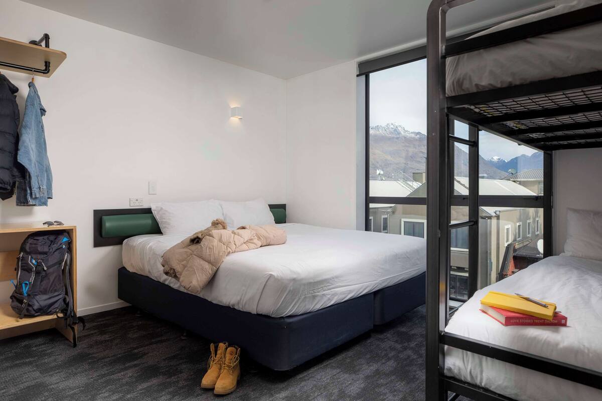 Ein Hotelzimmer mit Etagenbett, einem Einzelbett, einem kleinen Schreibtisch mit Stuhl, einem Rucksack und einem Paar Stiefel auf dem Boden.
