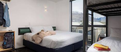 Une chambre d’hôtel avec un lit superposé, un lit simple, un petit bureau avec une chaise, un sac à dos et une paire de bottes posées sur le sol.