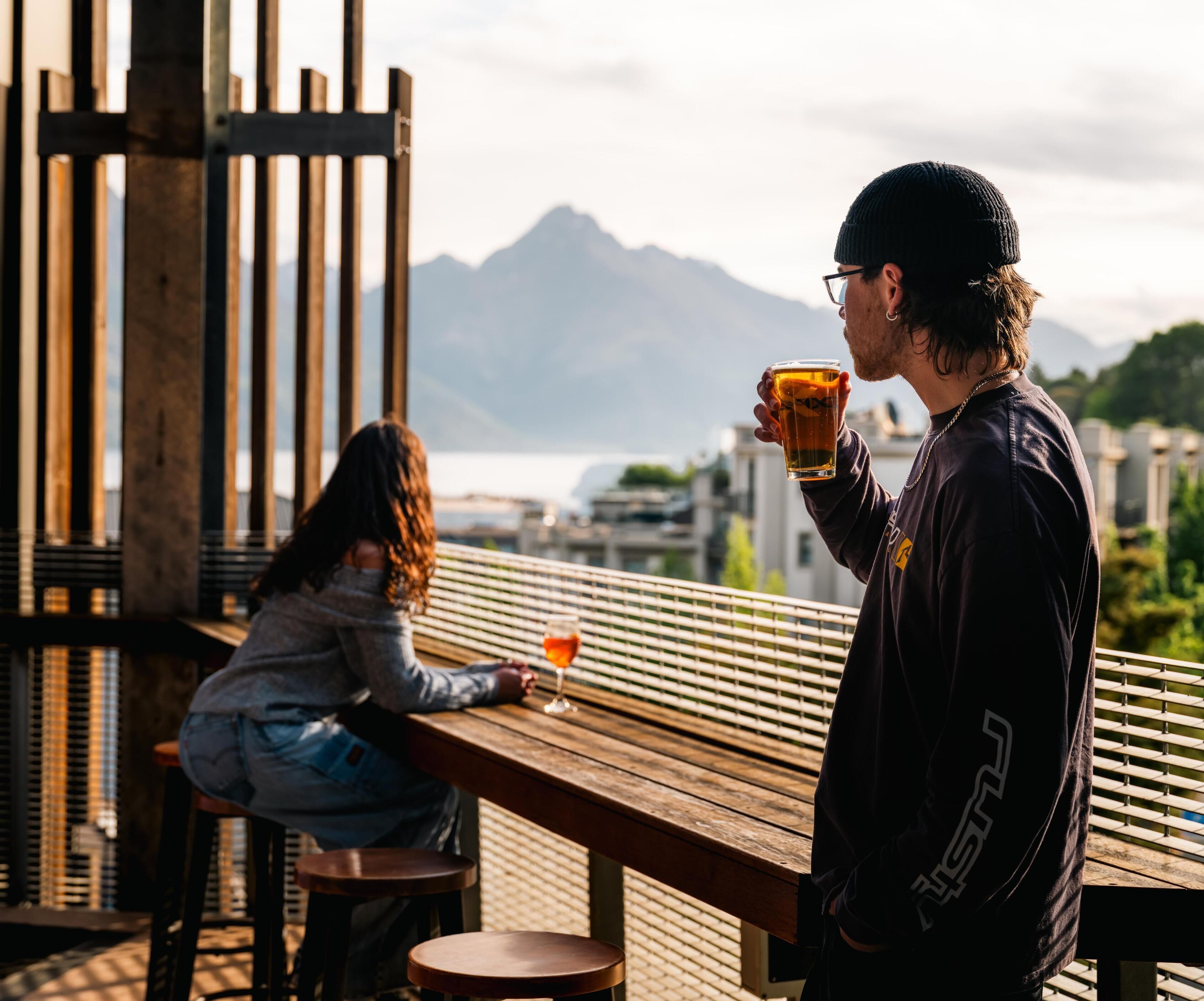 Deux personnes sont assises à une table en bois à l’extérieur, avec une vue sur les montagnes en arrière-plan.