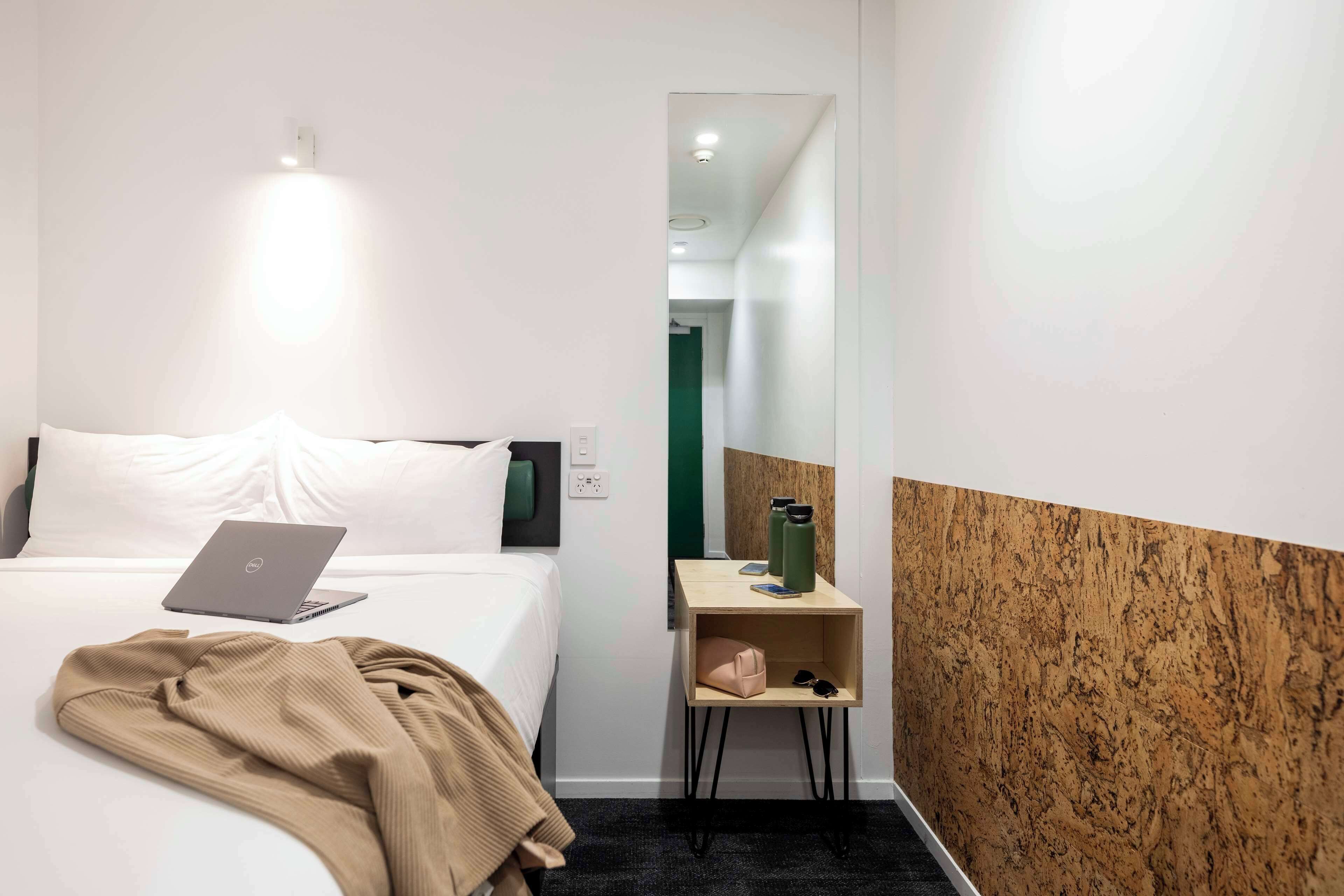 A hotel room with a bed, a laptop, a bedside table with a green water bottle, and a mirror.