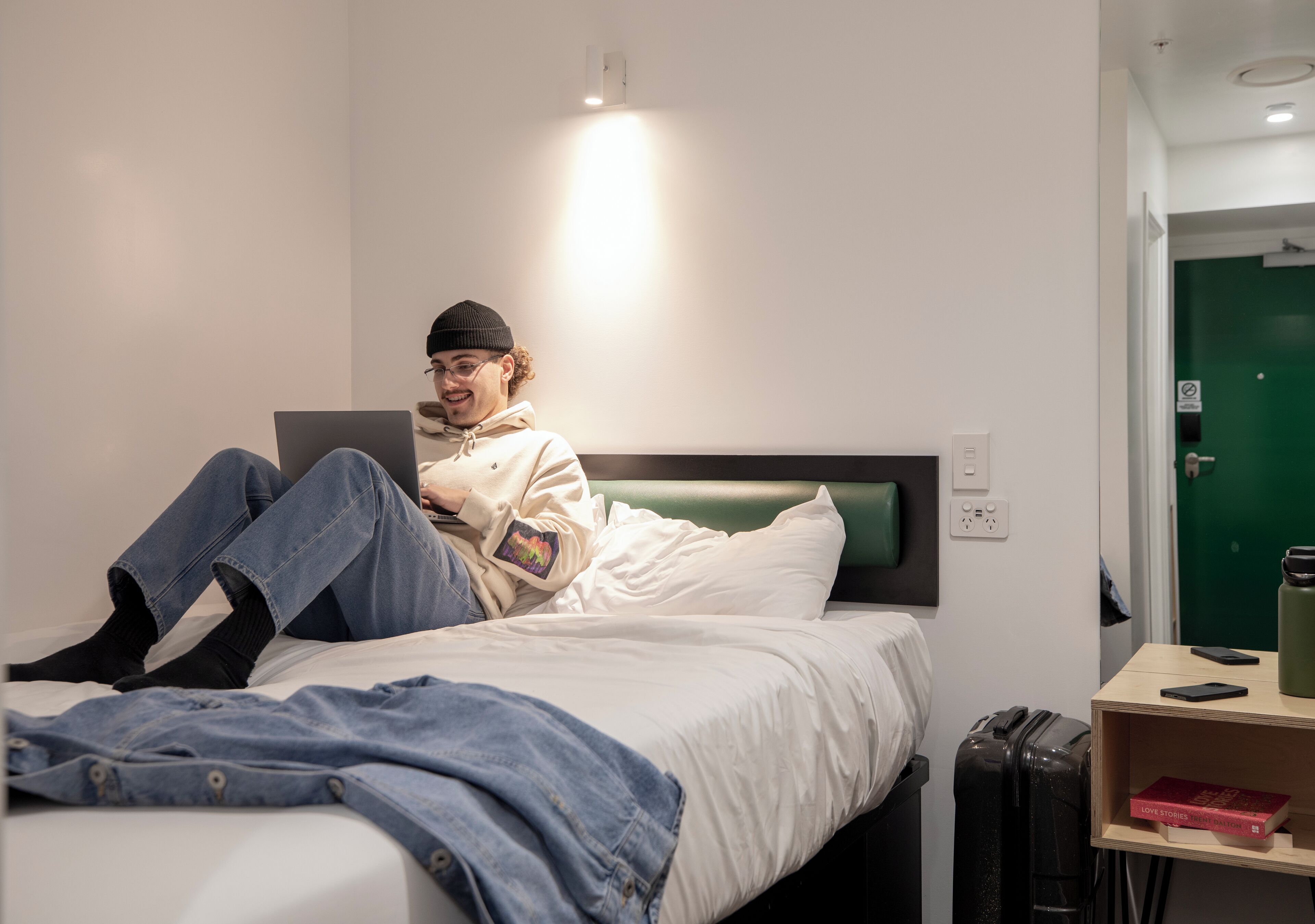 A person sitting on a bed in a room with a green door, a bedside table with a book, and a suitcase.