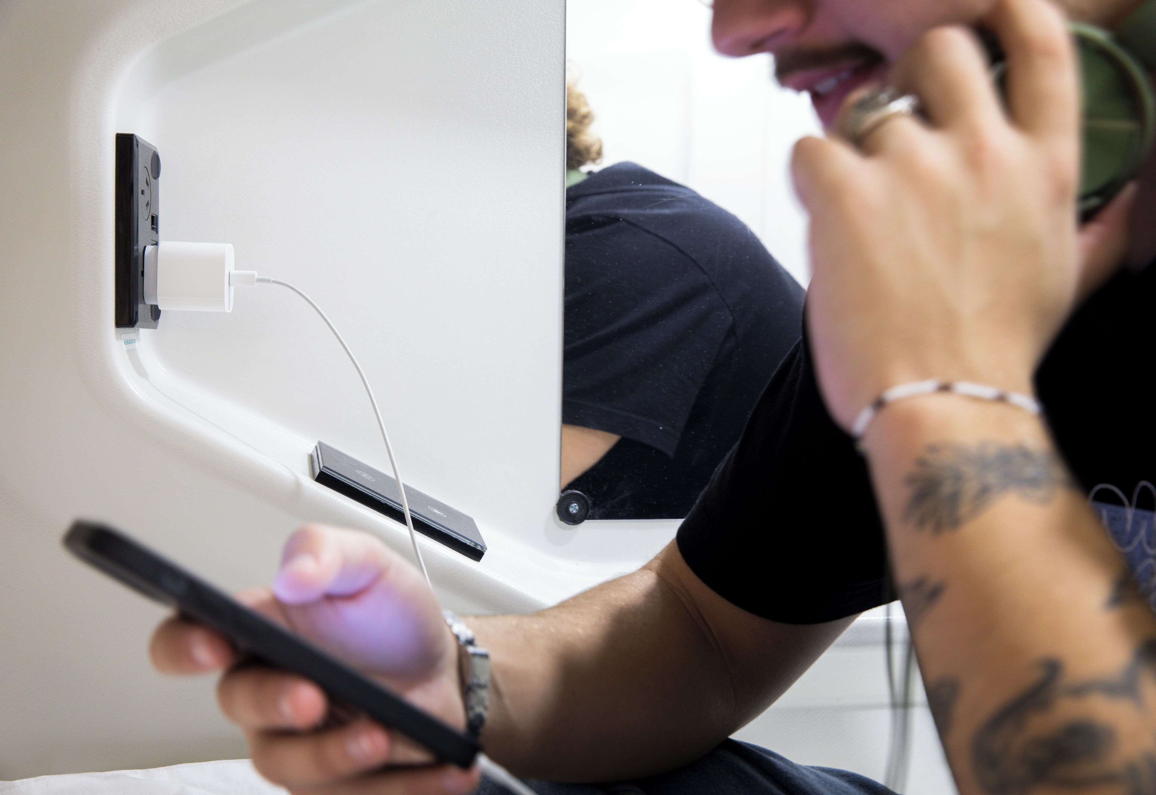A person sitting at a table with a laptop and a smartphone, a power outlet, and a wall-mounted device.
