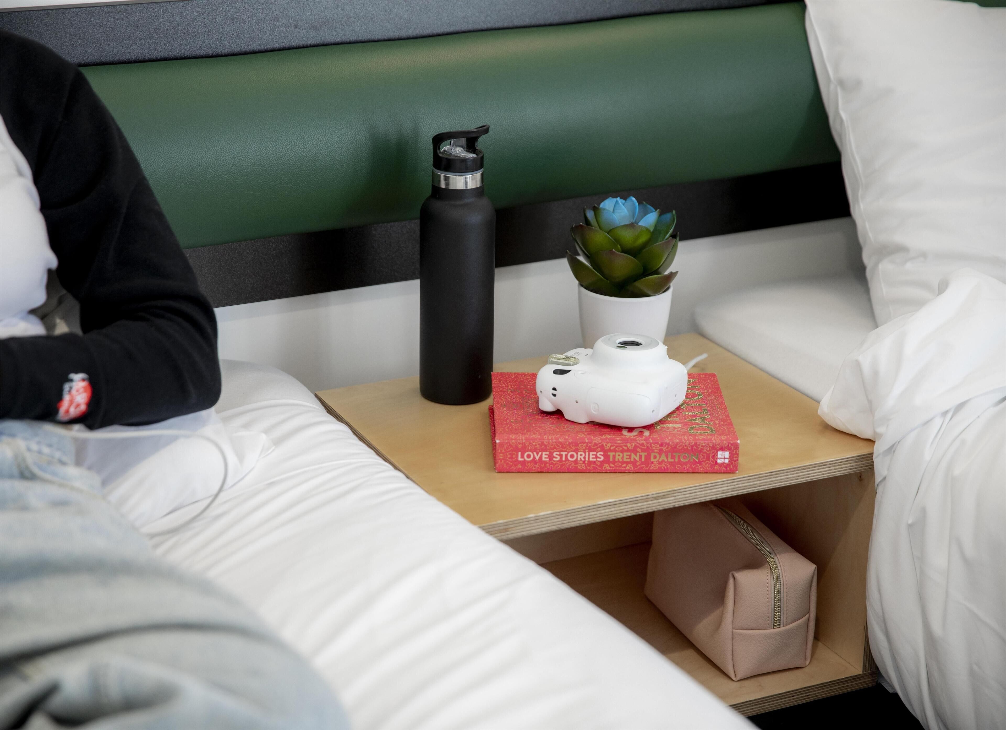 A neatly made bed with white linens, a black pillow, a black water bottle, a white camera on a red book titled 'LOVE STORIES', and a small potted plant.