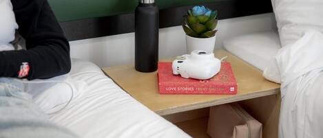 A neatly made bed with white linens, a black pillow, a black water bottle, a white camera on a red book titled 'LOVE STORIES', and a small potted plant.