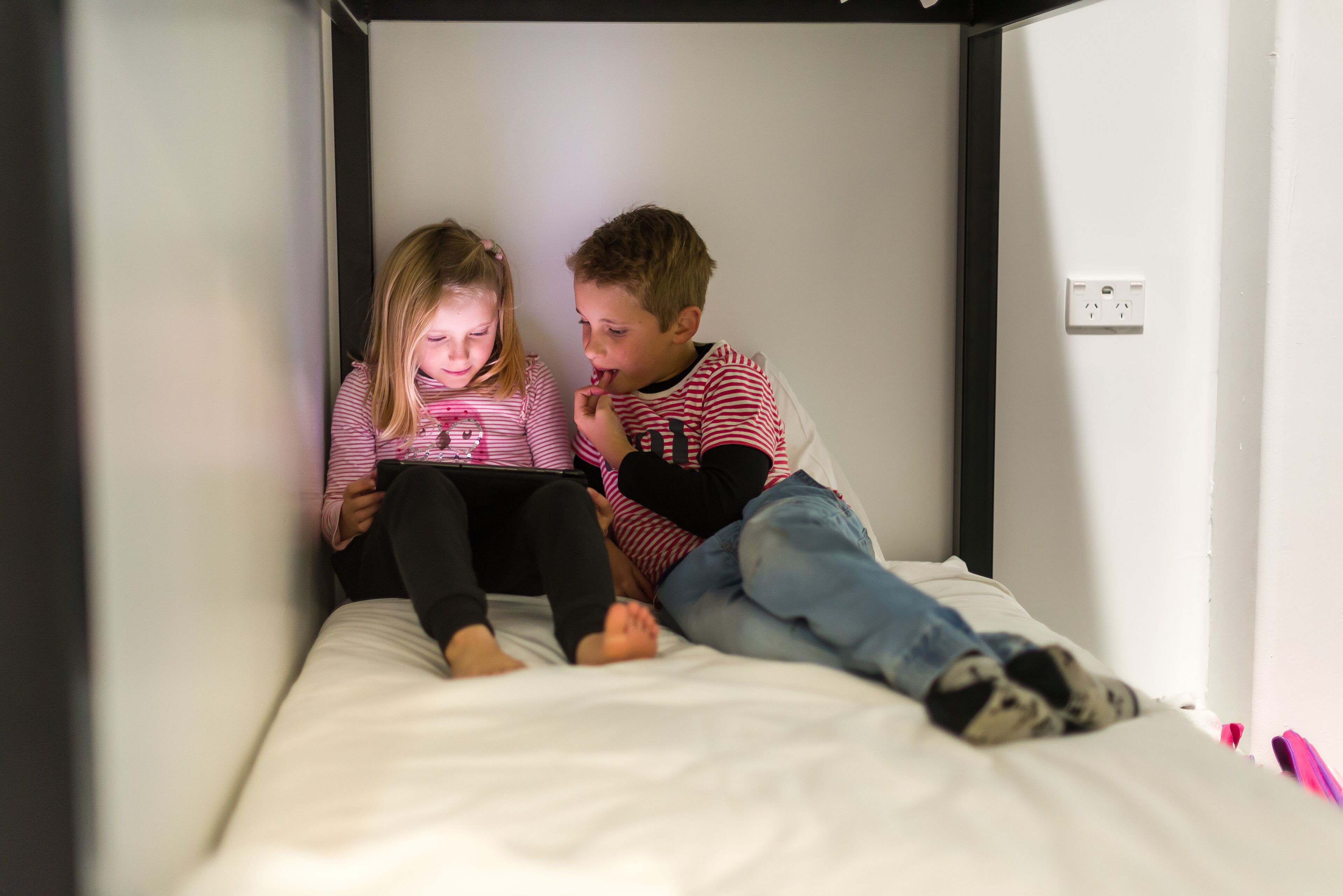 Dos niños sentados en una cama en una habitación con paredes blancas y un cabecero negro.